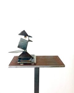 a metal sculpture of a chickadee made from scrap pieces of metal is sitting on a metal podium with a white background.