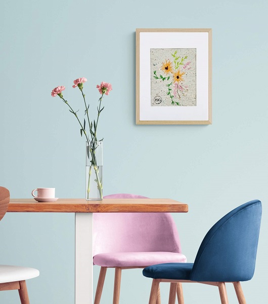 artwork of yellow and pink flowers embroidered on handmade paper displayed in a dinning room framed in a white frame against a blue wall