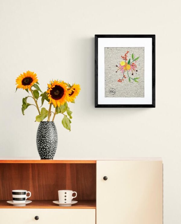 artwork for yellow flowers embroidered on handmade paper displayed in a black frame nest to a dresser with vase full of sunflowers